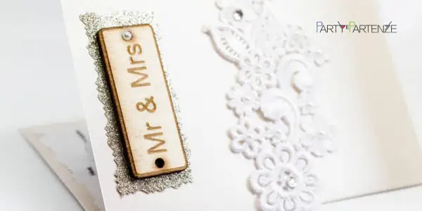 Dettagli di un invito di nozze: targa in legno "Mr & Mrs", pizzo bianco e logo "Party Partenze". Un esempio di diversi tipi di nozze.