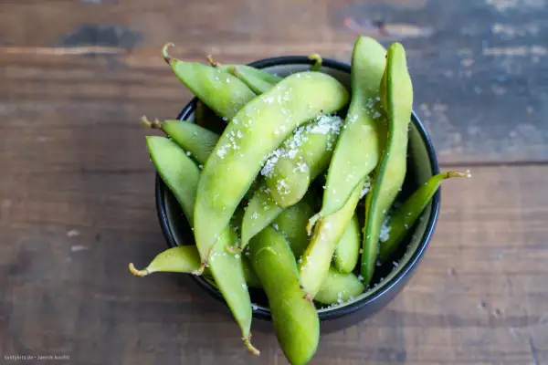 Grüne Edamame-Schoten, leicht gesalzen, in einer Schüssel auf Holzhintergrund. Was sind Edamame? Ein gesunder Snack!