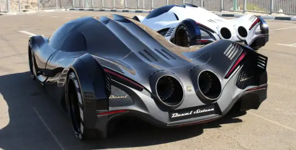 Dwa futurystyczne samochody Devel Sixteen, jeden czarny, drugi biały, stoją na asfalcie.