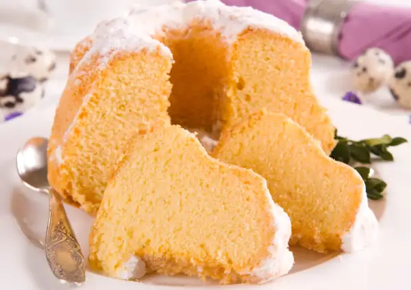 Puszysta babka z mascarpone, oprószona cukrem pudrem, z kilkoma kawałkami na talerzu. Obok leży łyżeczka.