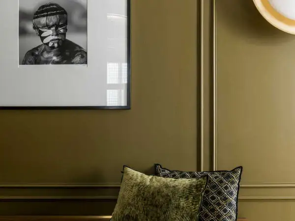 Un pan de mur vert olive, orné d'un portrait encadré d'un jeune homme au visage peint, et de coussins décoratifs.
