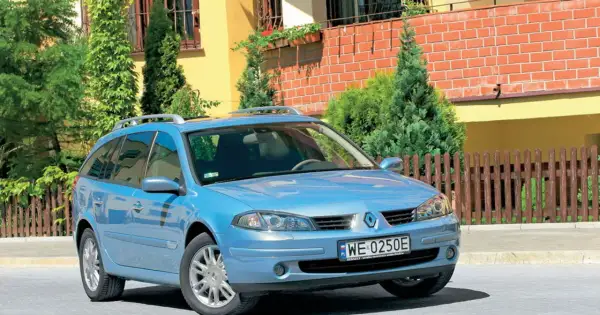 Błękitne kombi Renault Laguna 2 z relingami dachowymi stoi przed domem z żółtą elewacją i drewnianym płotem.