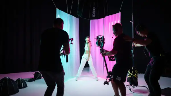 Filming a music video, learning how to color lighting with a dancer posed between blue and pink backdrops, while camera operators film.