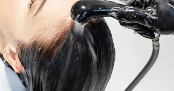Kobieta z ciemnymi włosami poddaje się zabiegowi **reverse washing**. Woda spływa po jej głowie, a dłoń w czarnej rękawiczce trzyma słuchawkę prysznicową.
