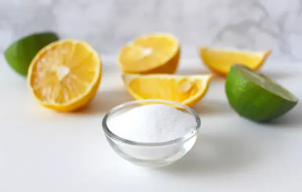 Una ciotola di cristalli bianchi, che ricordano la struttura dell'acido citrico, circondata da spicchi di limone e lime freschi.