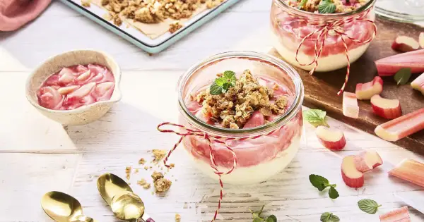 Zwei Gläser mit Rhabarber Crumble, garniert mit Minze. Frischer Rhabarber liegt daneben.