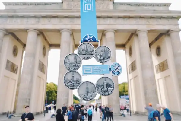 Medal za ukończenie korony maratonów świata przed Bramą Brandenburską. Widoczne miasta: Boston, Tokio, Nowy Jork, Chicago, Berlin, Londyn.