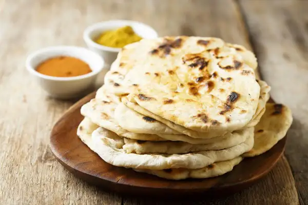 Pila di pane naan appena sfornato, con due ciotoline di spezie in sottofondo.