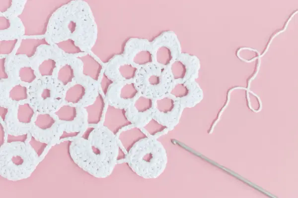 White Irish crochet lace with flowers and hearts, a crochet hook, and white yarn on a pink background.