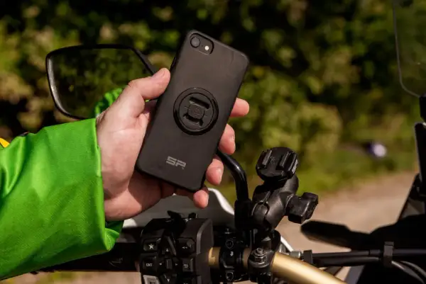 Dłoń trzyma czarne etui na telefon z mocowaniem SP Connect, obok uchwytu na kierownicy motocykla.