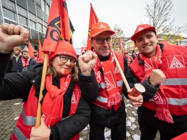 Drei Personen in roten Westen und Schals mit Gewerkschaftssymbolen, die Fahnen schwenken und die Faust ballen.