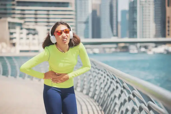 Une coureuse en tenue de sport, écouteurs aux oreilles, souffre d'un point de côté aigu en pleine course le long d'une promenade urbaine.