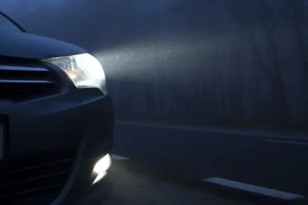 El coche avanza en la niebla con sus luces antiniebla encendidas, iluminando el camino oscuro y los árboles a un lado.