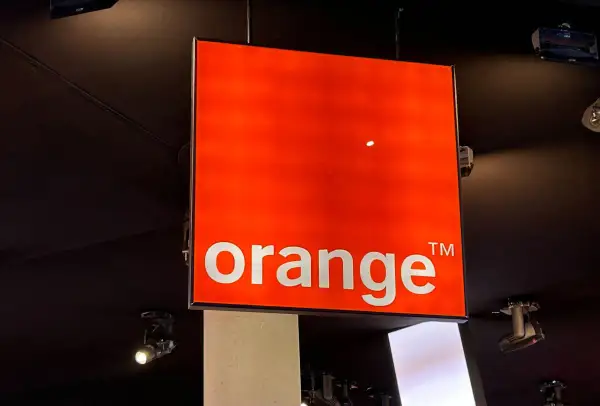 Jasny, pomarańczowy znak z napisem "orange" wisi pod sufitem. To logo Orange, które często pojawia się na ekranach telewizorów.
