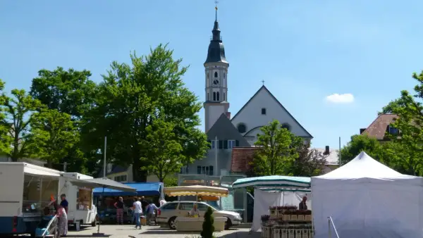 Bunter Markt in Mering mit Kirche im Hintergrund. Stände mit Blumen und Lebensmitteln, Menschen bummeln.