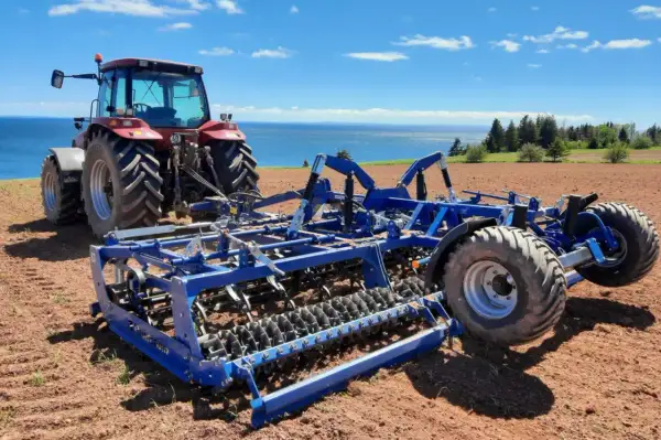 Czerwony traktor z niebieskim agregatem uprawowym na polu, gotowy do siewu. W tle błękitne niebo i morze.