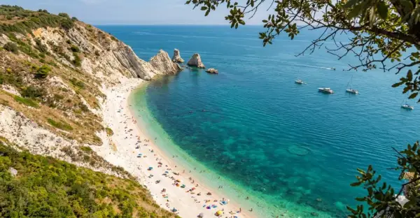 Spiaggia affollata ai piedi del Monte Conero, con acque cristalline e barche all'orizzonte.