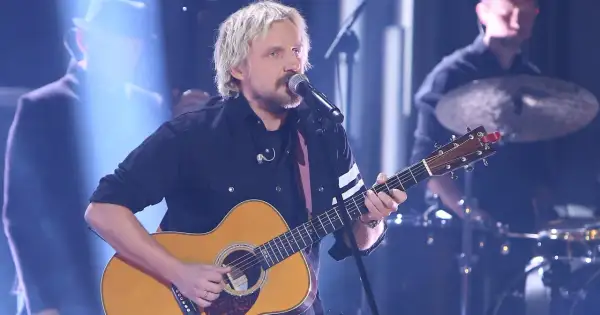 Artysta z blond włosami gra na gitarze akustycznej, śpiewając do mikrofonu. Juz teraz wiem ze dni tekst.