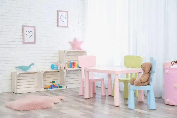 Kolorowy stolik z krzesełkami dla dzieci, idealny do zabawy i nauki. Na ścianie wiszą ramki z serduszkami, a obok stoją drewniane skrzynki z zabawkami.