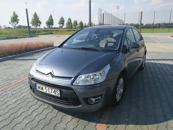 Szary Citroen C4 z 2008 roku na parkingu z kostki brukowej, w tle drzewa i boisko.