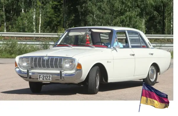 Vit Ford Taunus, en klassisk modell av svenska bilar, parkerad utomhus med en tysk flagga i förgrunden.