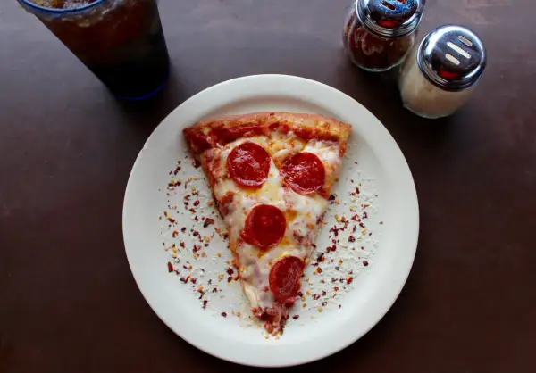 Kawałek pizzy pepperoni na talerzu, obok słoiczki z przyprawami i napój. Zastanawiasz się, ile kosztuje pizza we Włoszech?