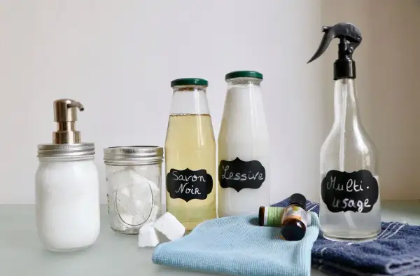 Bouteilles étiquetées "Savon Noir", "Lessive" et "Multi-usage", ainsi qu'un distributeur de savon, des pastilles et des huiles essentielles. Des produits faits maison pour un nettoyage écologique.