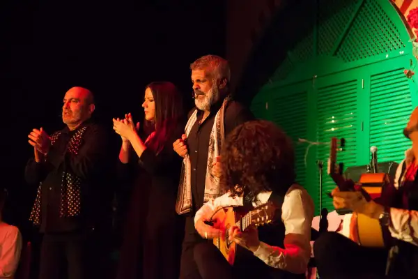 Artistas flamencos, incluyendo cantaores flamencos famosos, aplauden y tocan la guitarra en un escenario con una puerta verde de fondo.
