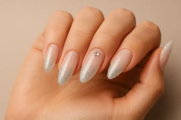 Main d'une femme aux ongles en amande, ornés d'un dégradé de paillettes argentées et d'un petit strass sur l'annulaire.