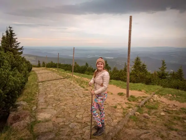 Uśmiechnięta turystka z kijkami na kamienistej ścieżce. Widok na góry i doliny. Czas wejścia na Śnieżkę zależy od kondycji i wybranej trasy.