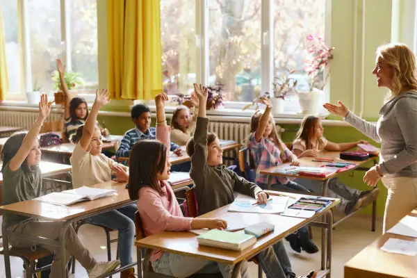 Grundschüler mit erhobenen Händen, bereit zu antworten. Die Sitzordnung zur Wand hin ist gut erkennbar, die Lehrerin lächelt.