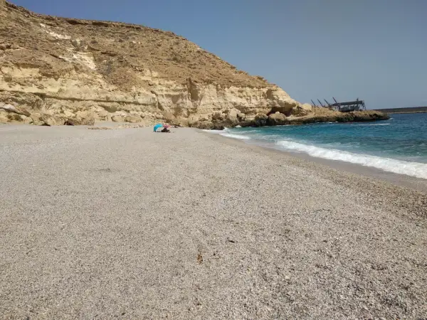 Arena y guijarros en la playa de las salinicas, con acantilados dorados y mar azul. Una sombrilla azul protege a unos bañistas.