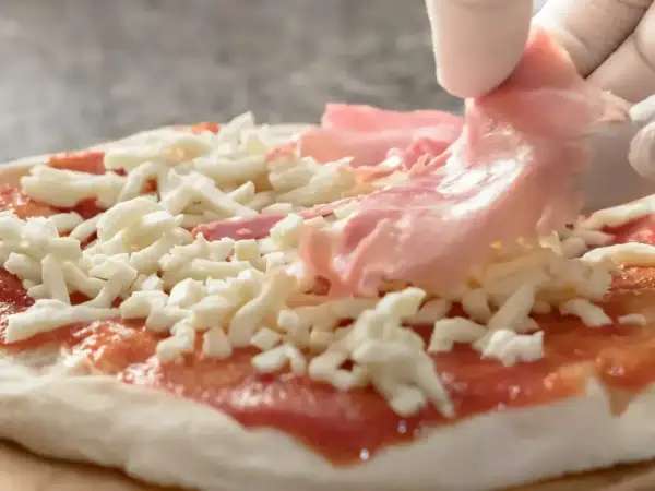 Rękawiczka w białej rękawiczce dodaje cienkie plastry różowej szynki na pizzę, która jest już pokryta sosem pomidorowym i startym serem.
