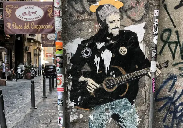 Graffiti di Pino Daniele con la chitarra, un omaggio a Napoli. I testi "Resta... resta cu'mmè" risuonano tra i vicoli.