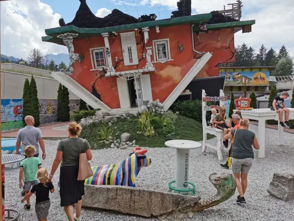 Ein rotes Haus steht auf dem Kopf, die Treppe führt ins Nichts. Eine Familie bestaunt das Spektakel, bunte Skulpturen und ein Dino-Land im Hintergrund.