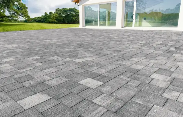 Un ampio patio in gres carrabile grigio, con pavimentazione a motivi geometrici, si estende davanti a una moderna casa con vetrate.