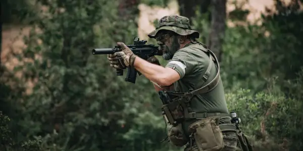 Jugador de deporte airsoft con camuflaje facial y equipo táctico, apuntando con su réplica de rifle en un bosque.