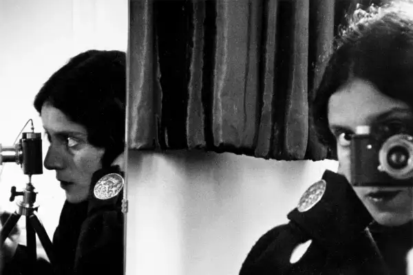 Tina Modotti w kadrze, z aparatem w ręku, patrzy w obiektyw.