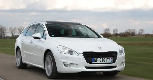 Biały Peugeot 508 SW z czarnym dachem jedzie po drodze, otoczony zielonymi polami i drzewami.