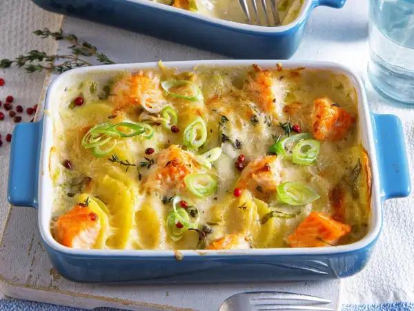 Herzhaft überbackener Lachs mit Kartoffeln in einer blauen Auflaufform, garniert mit Frühlingszwiebeln und rosa Pfefferbeeren.
