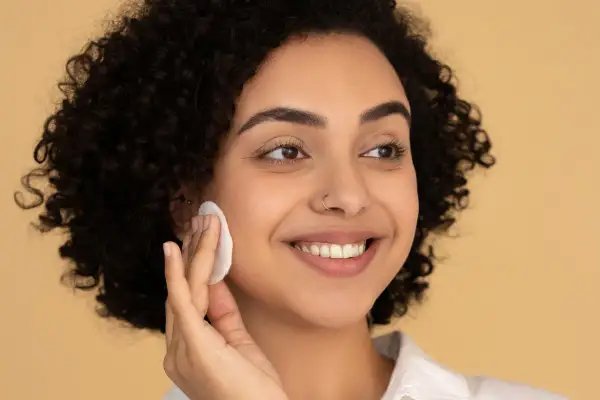 Donna sorridente con capelli ricci usa un dischetto per struccarsi, mostrando come si usa il burro struccante per una pelle luminosa.