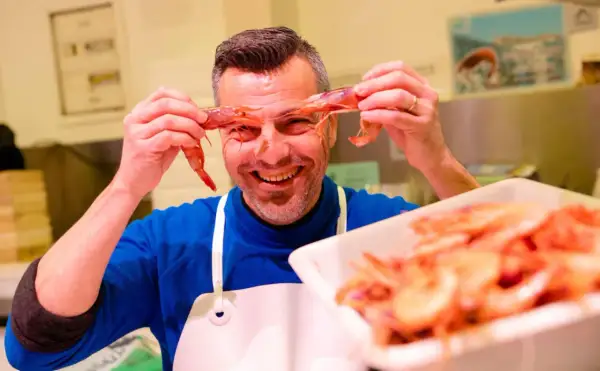 Hombre sonriente jugando con mariscos rojos, sosteniendo dos gambas frente a sus ojos.
