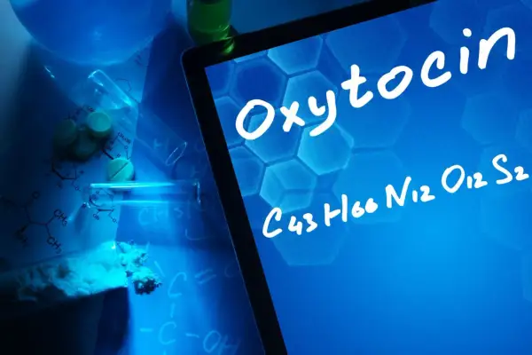 Na ekranie tabletu widnieje napis "Oksytocyna co to" i jej wzór chemiczny. W tle leżą tabletki, proszek i probówki.