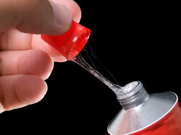 Main d'une personne tenant le bouchon rouge d'un tube de colle forte, avec des fils de colle s'étirant. Astuce pour comment enlever de la colle forte sur du métal.