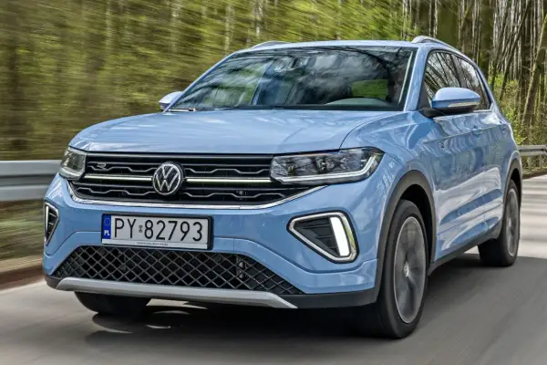Niebieski VW T-Cross pędzi leśną drogą, odbijając drzewa w szybie.