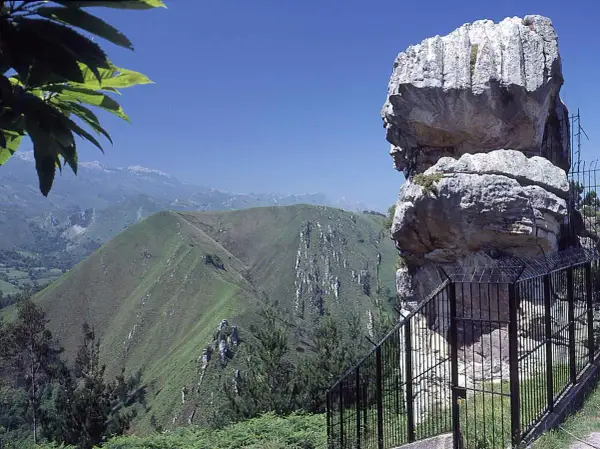 El idolo de peña tu, una roca con forma de perfil humano, vigila un paisaje montañoso verde y sereno bajo un cielo azul.