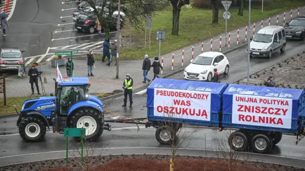 Niebieski traktor ciągnie przyczepę z banerami "Pseudoekolodzy was oszukują" i "Unijna polityka zniszczy rolnika".
