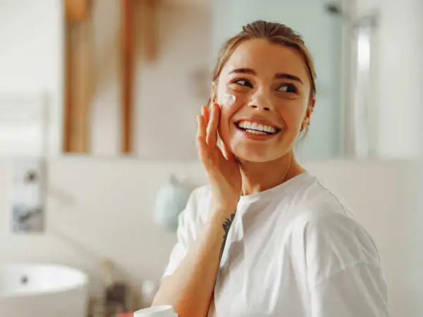 Une femme souriante applique une crème sur sa joue. Elle se prépare pour la journée, peut-être un primer avant ou après crème solaire.