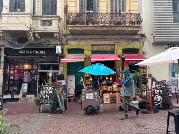Un hombre con capucha camina por una calle adoquinada en Montevideo, pasando por tiendas de discos y un puesto de frutas.