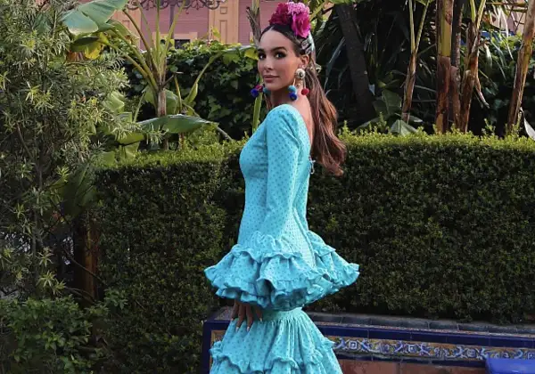 Una mujer luce un vestido de flamenca turquesa con lunares, adornado con volantes y una flor en la cabeza.